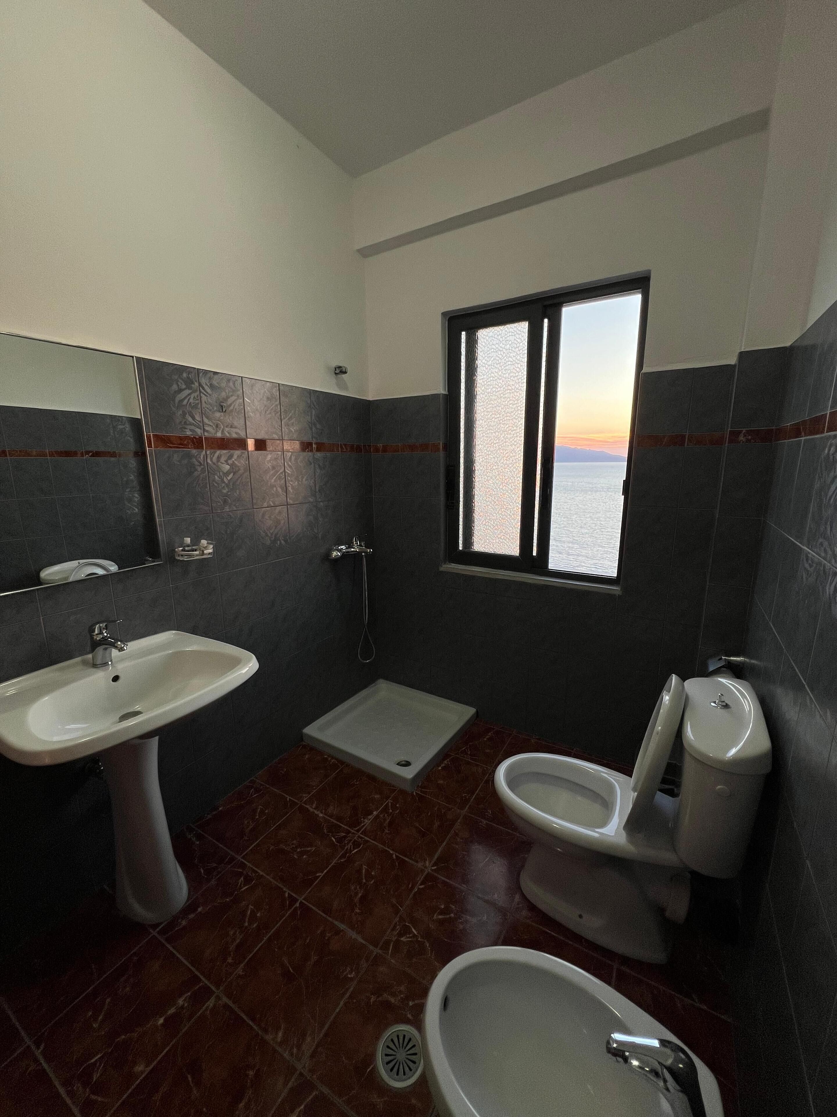 A bathroom with a sink, toilet, and shower area, featuring dark tiles and a window with a view.