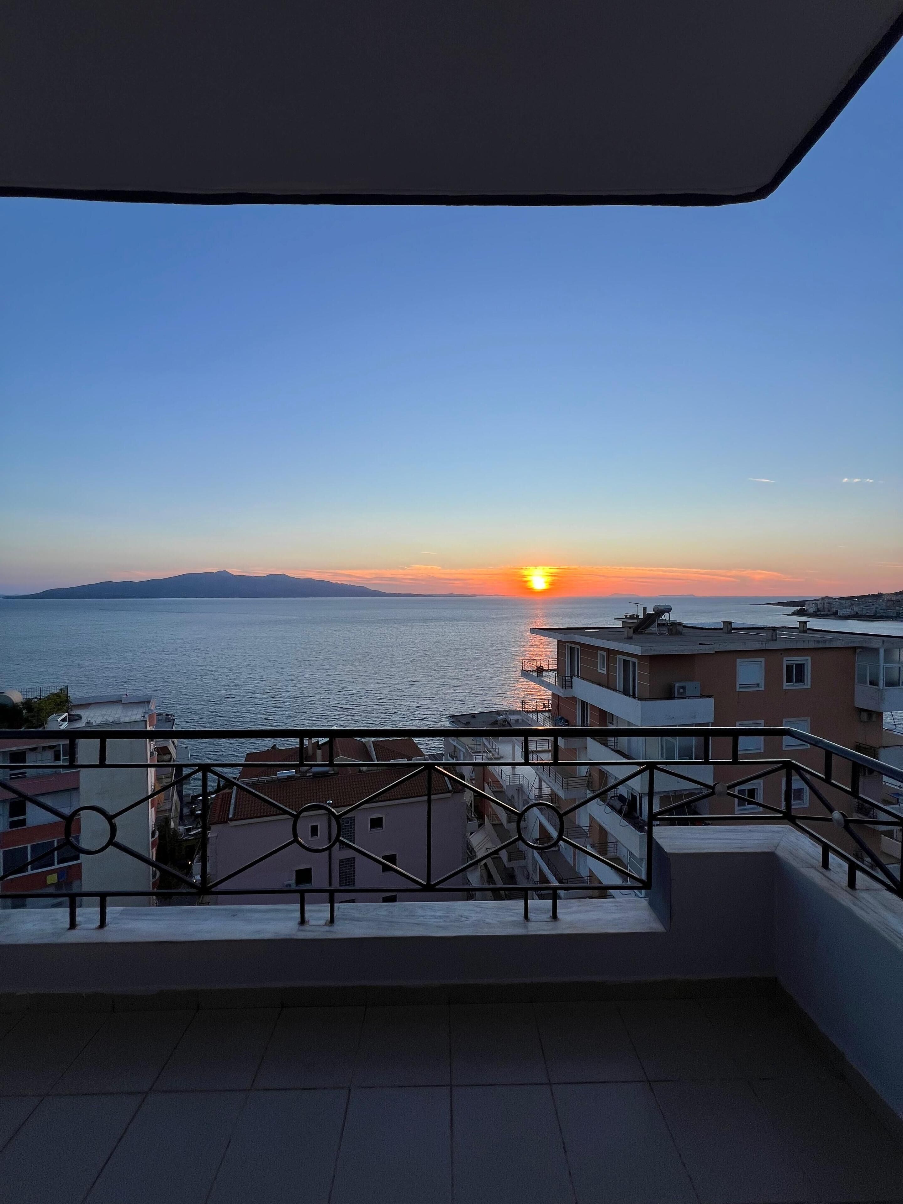 A balcony with a view of the sunset over the sea, buildings in the background, and a railing.