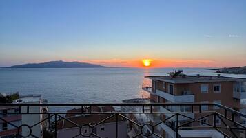 A balcony with a view of the sunset over the sea, buildings in the background, and a railing.