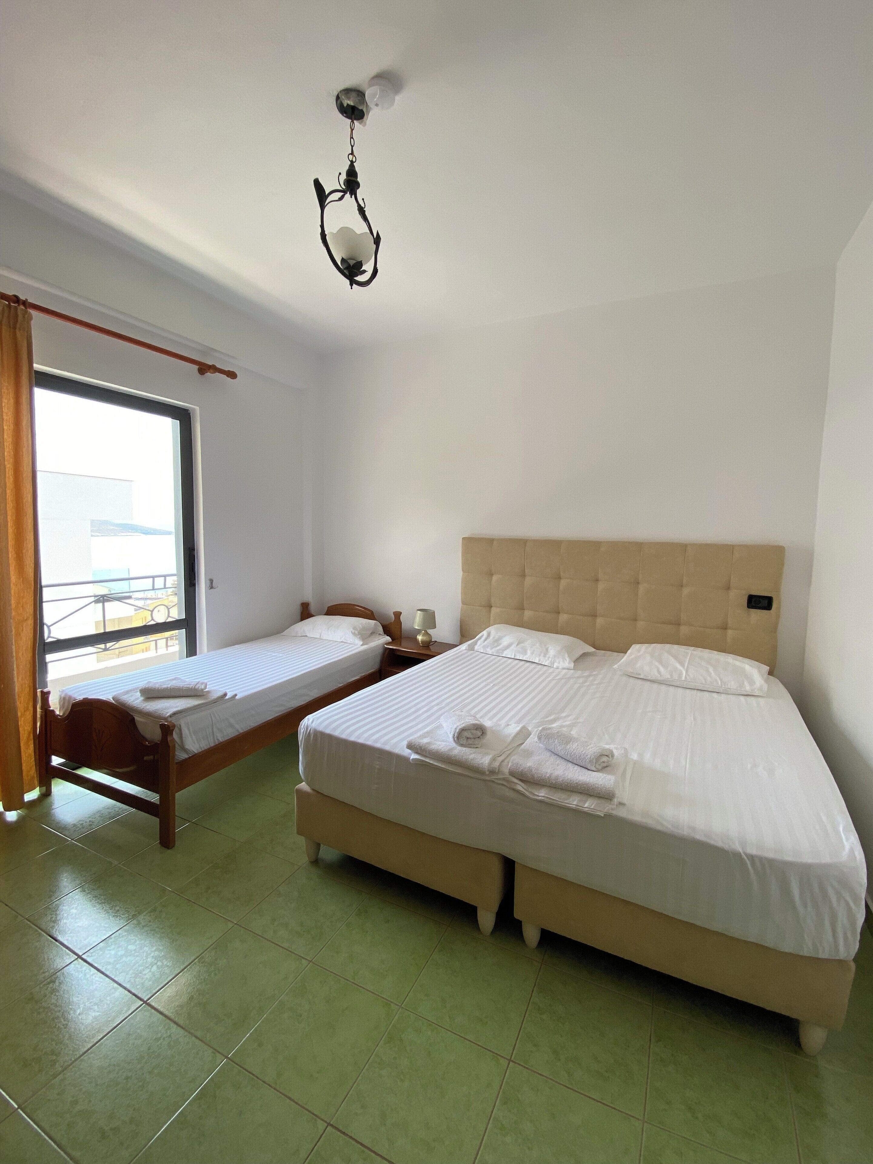 Ein Hotelzimmer mit zwei Einzelbetten, einem großen Fenster mit Vorhängen, einem hölzernen Kopfteil und einer Pendelleuchte.