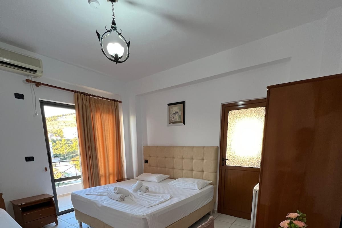 A hotel room with a bed, a dining table with chairs, a vase with flowers, a ceiling fan, and a window with curtains.