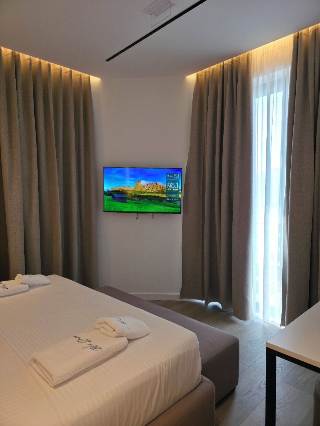 Una habitación de hotel con cama, televisor montado en la pared y cortinas en las ventanas.