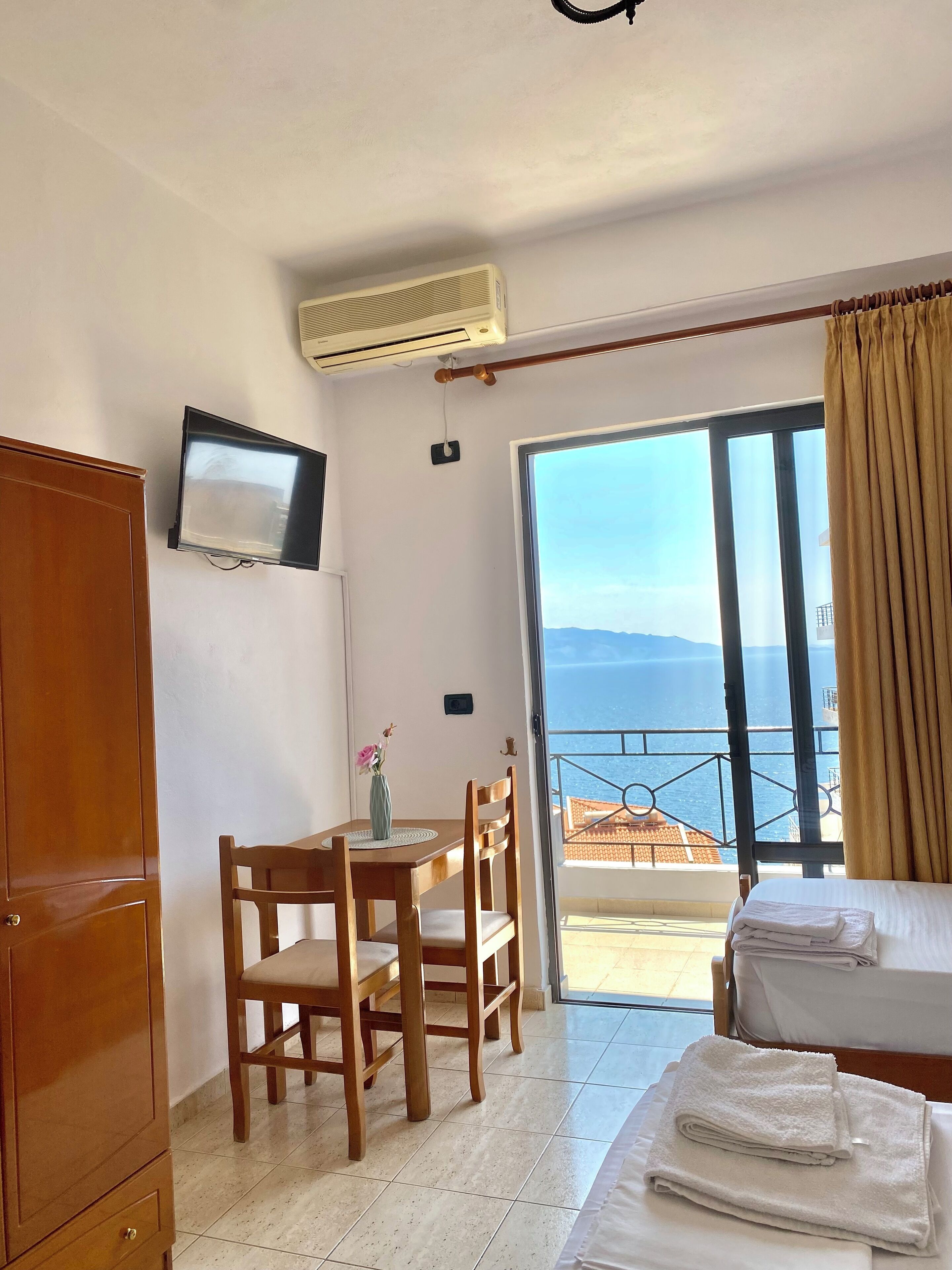 A hotel room with a bed, a desk with a chair, a TV mounted on the wall, and a balcony with a view of the sea.