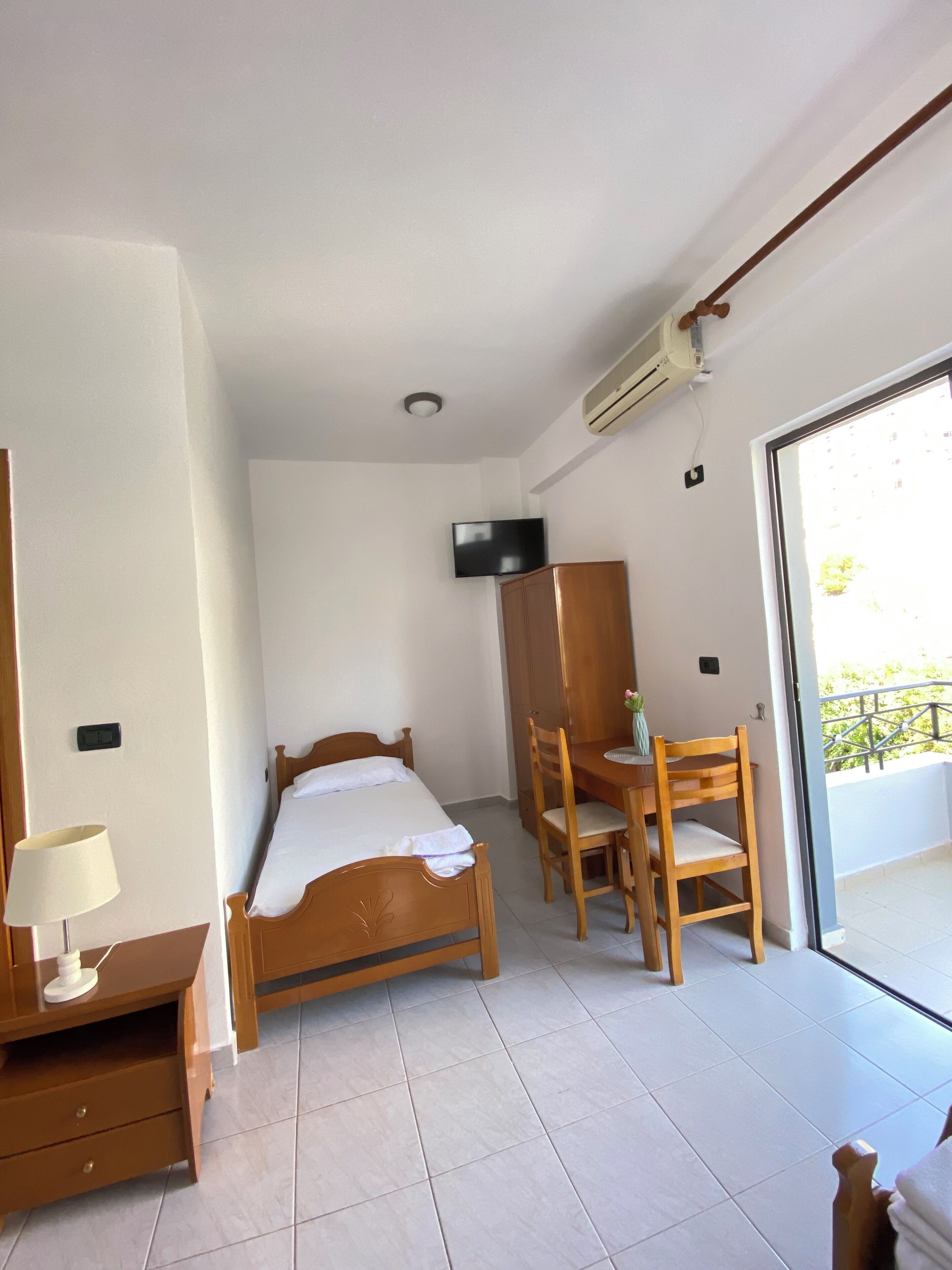 A bedroom with a wooden bed, a desk with a lamp, a TV mounted on the wall, and a balcony with a railing.
