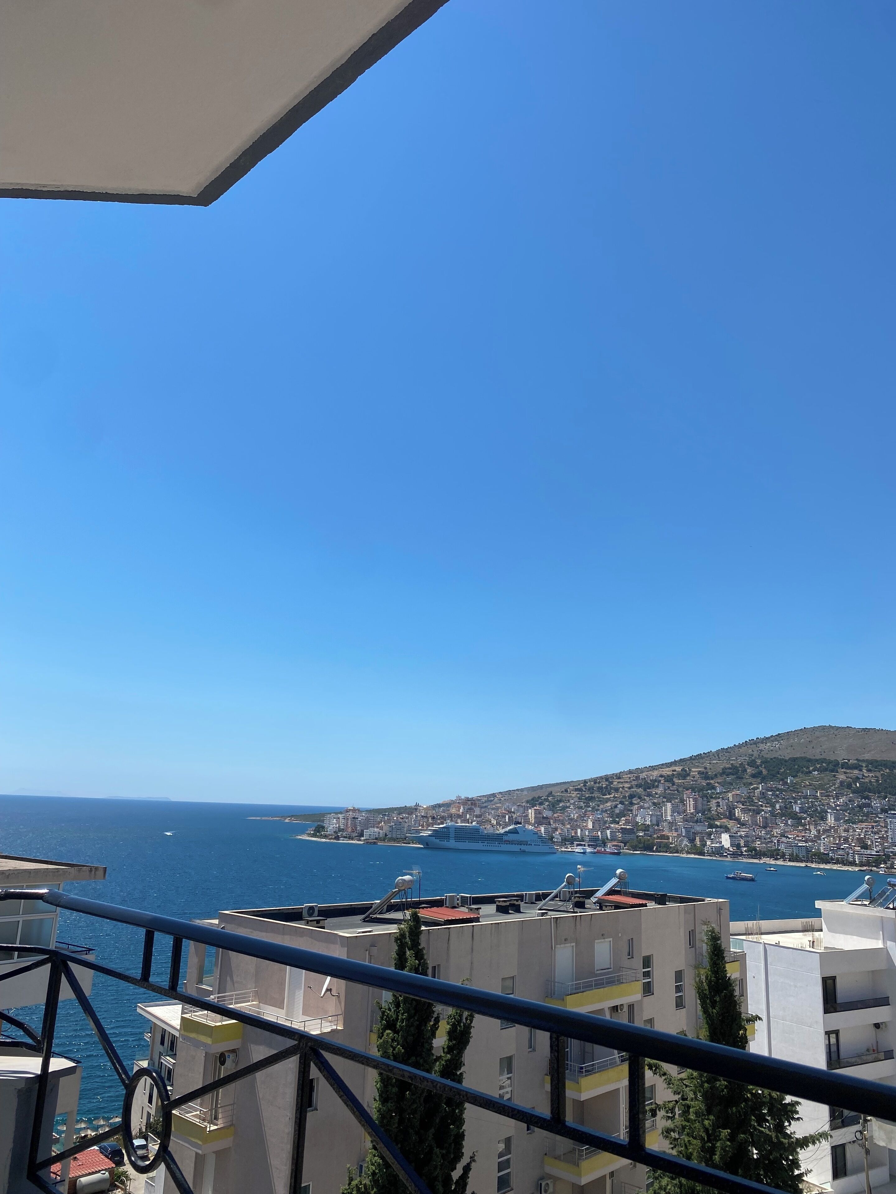 Een balkon met een helder uitzicht op zee, gebouwen en een heuvel in de verte.