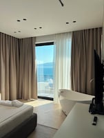 A modern hotel room with a large bed, a white desk, a bathtub, and a balcony with a view of the sea.