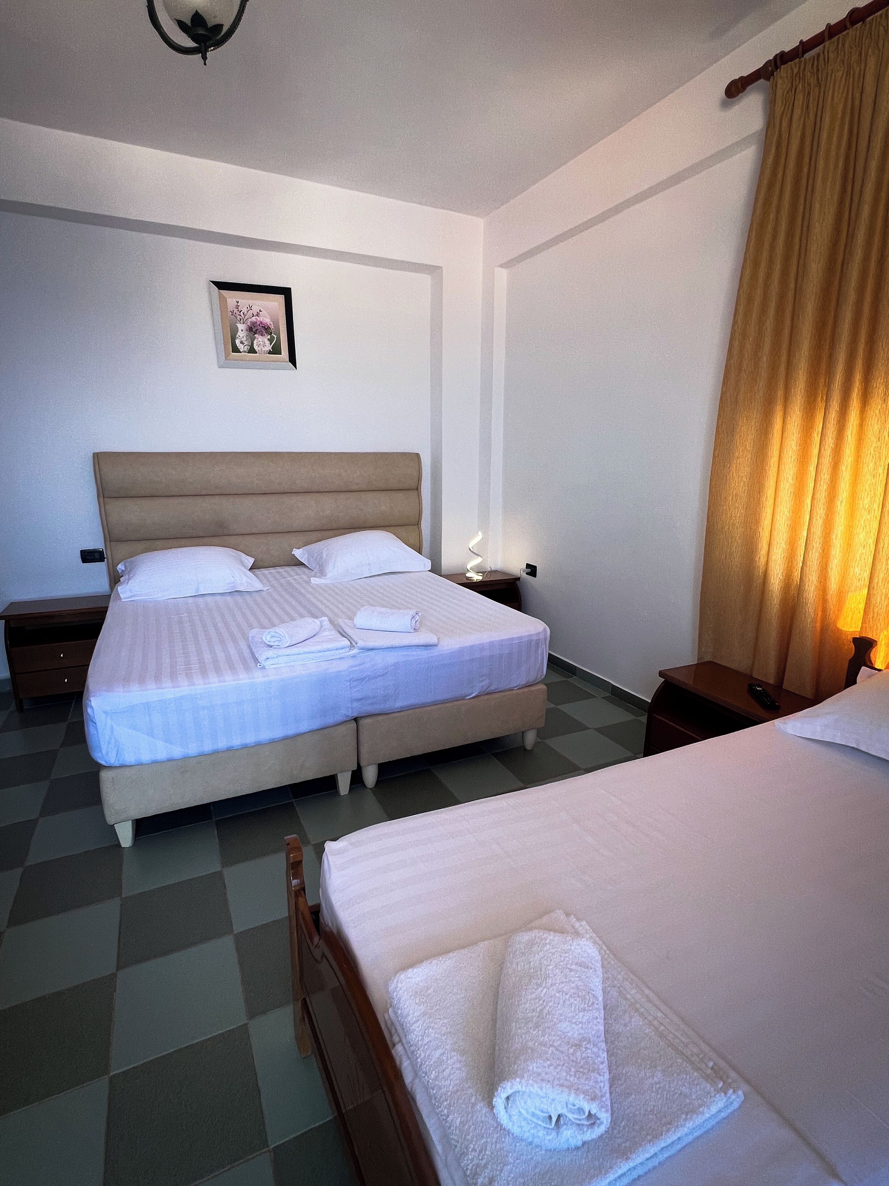 A hotel room with two beds, a nightstand with a lamp, a framed picture on the wall, and a checkered floor.