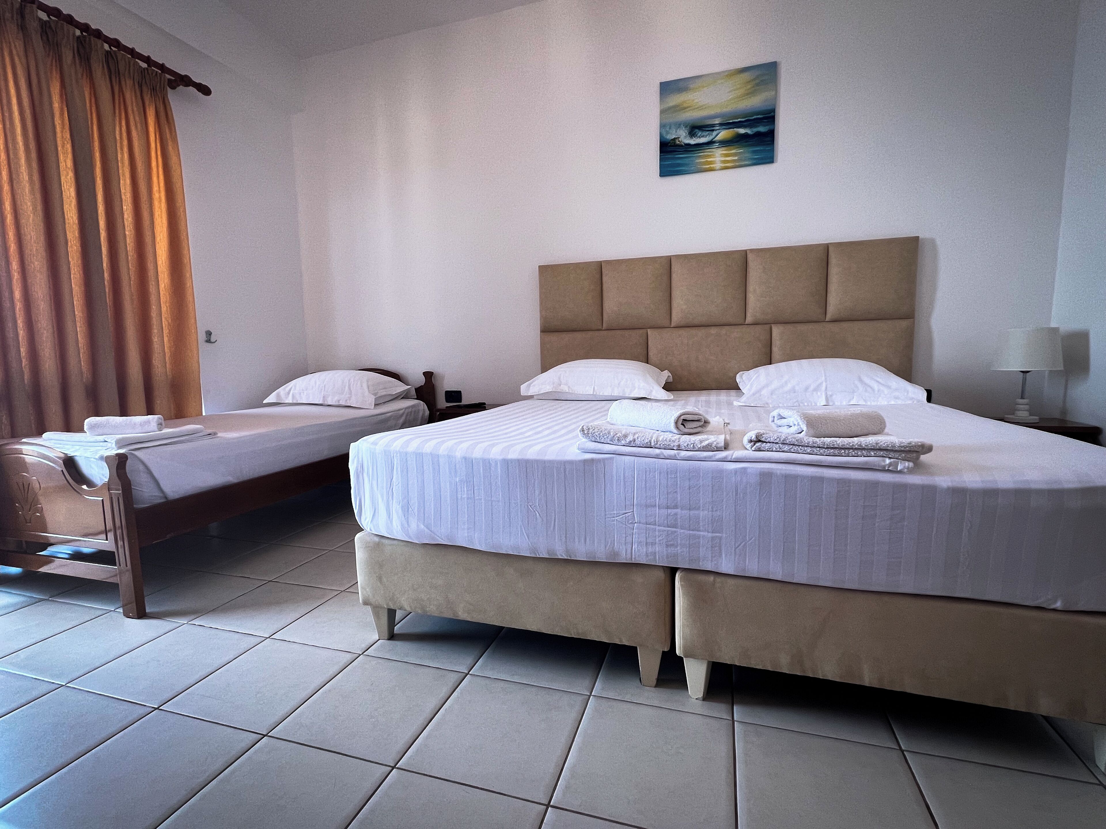 A hotel room with a large bed, two smaller beds, a nightstand, a lamp, and a painting on the wall.