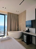 A modern hotel room with a large bathtub, a flat-screen TV, and a balcony with a view of the sea.