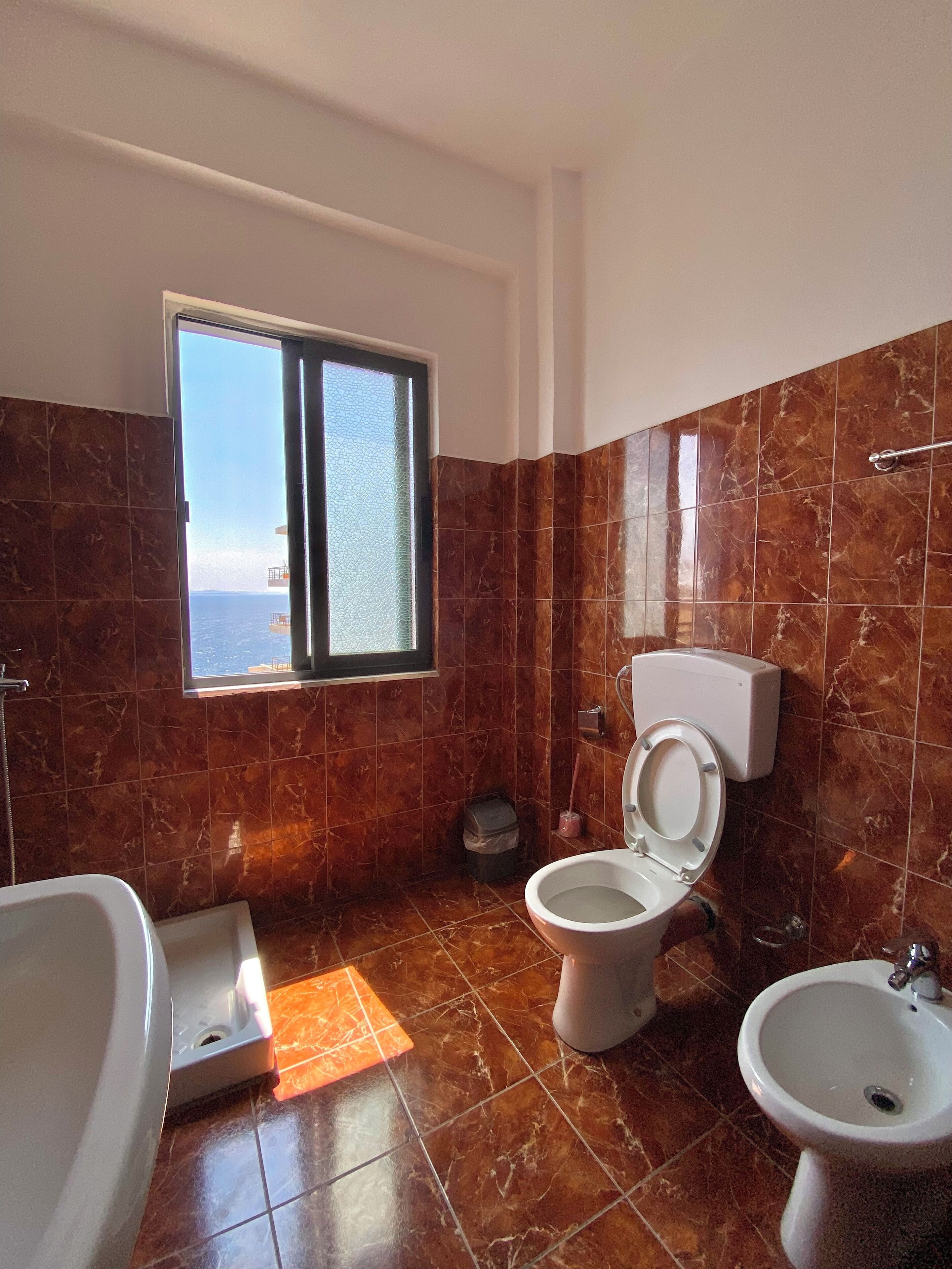 A bathroom with a toilet, bidet, and bathtub, featuring marble tiles and a window with a view.