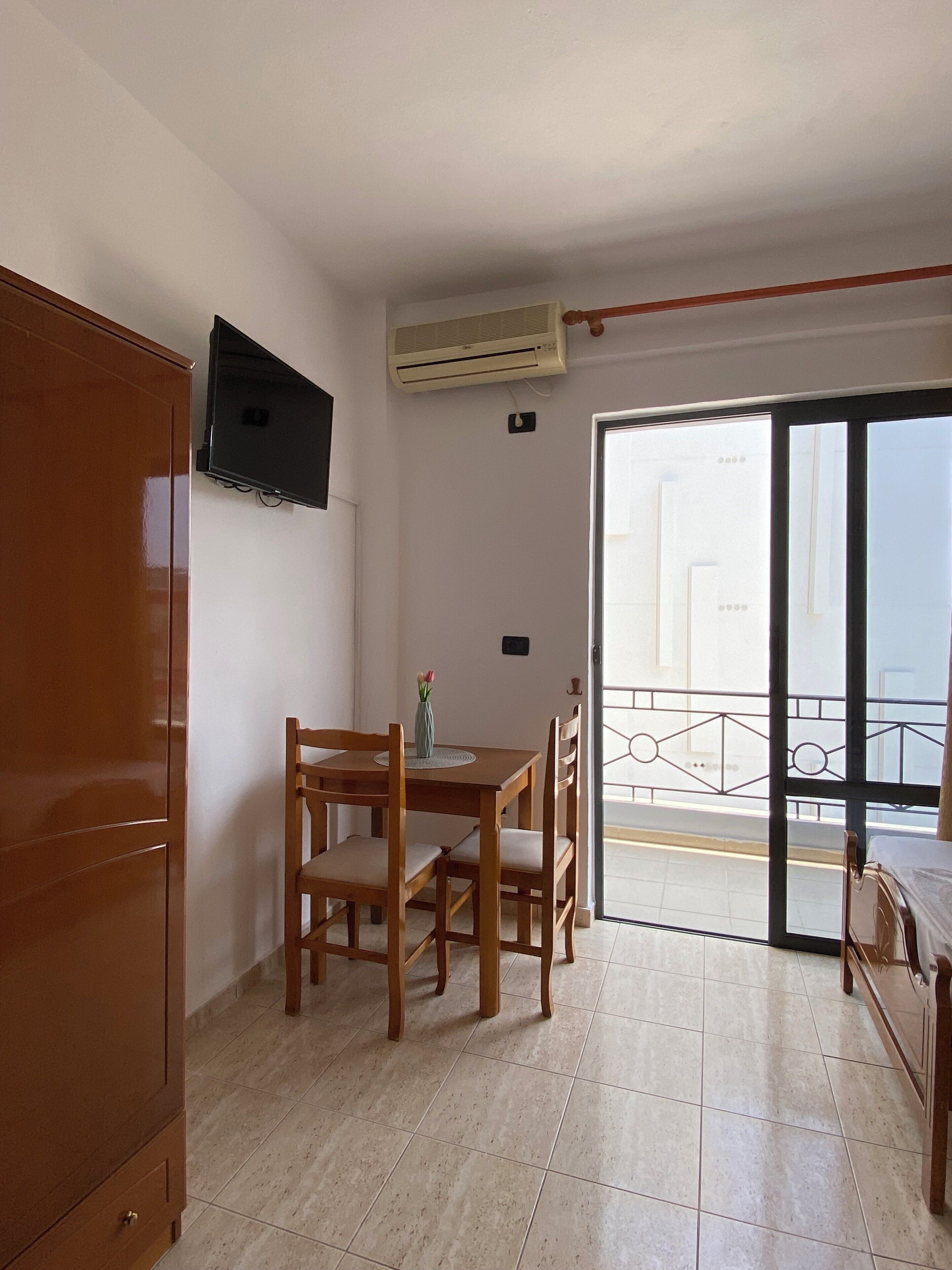 Una habitación con una mesa y sillas de madera, un televisor montado en la pared, un equipo de aire acondicionado y una puerta corrediza de vidrio que da a un espacio al aire libre.