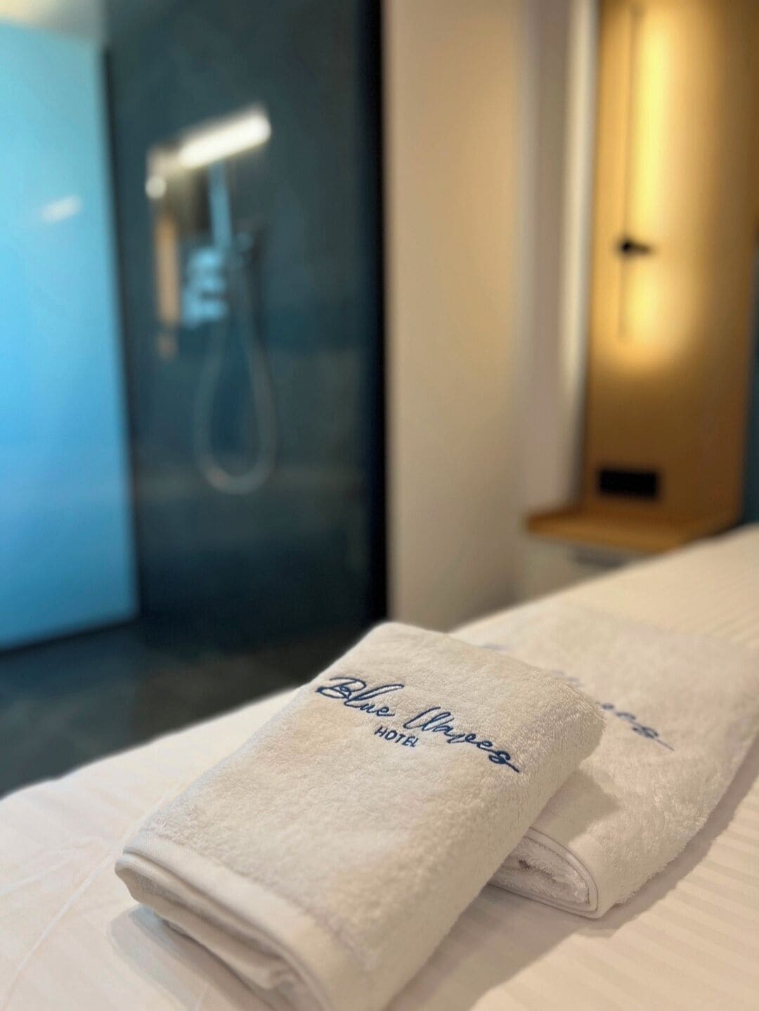 Ein ordentlich gefaltetes weißes Handtuch mit "Blue Waves Hotel" aufgestickt, platziert auf einem Bett in einem Hotelzimmer.