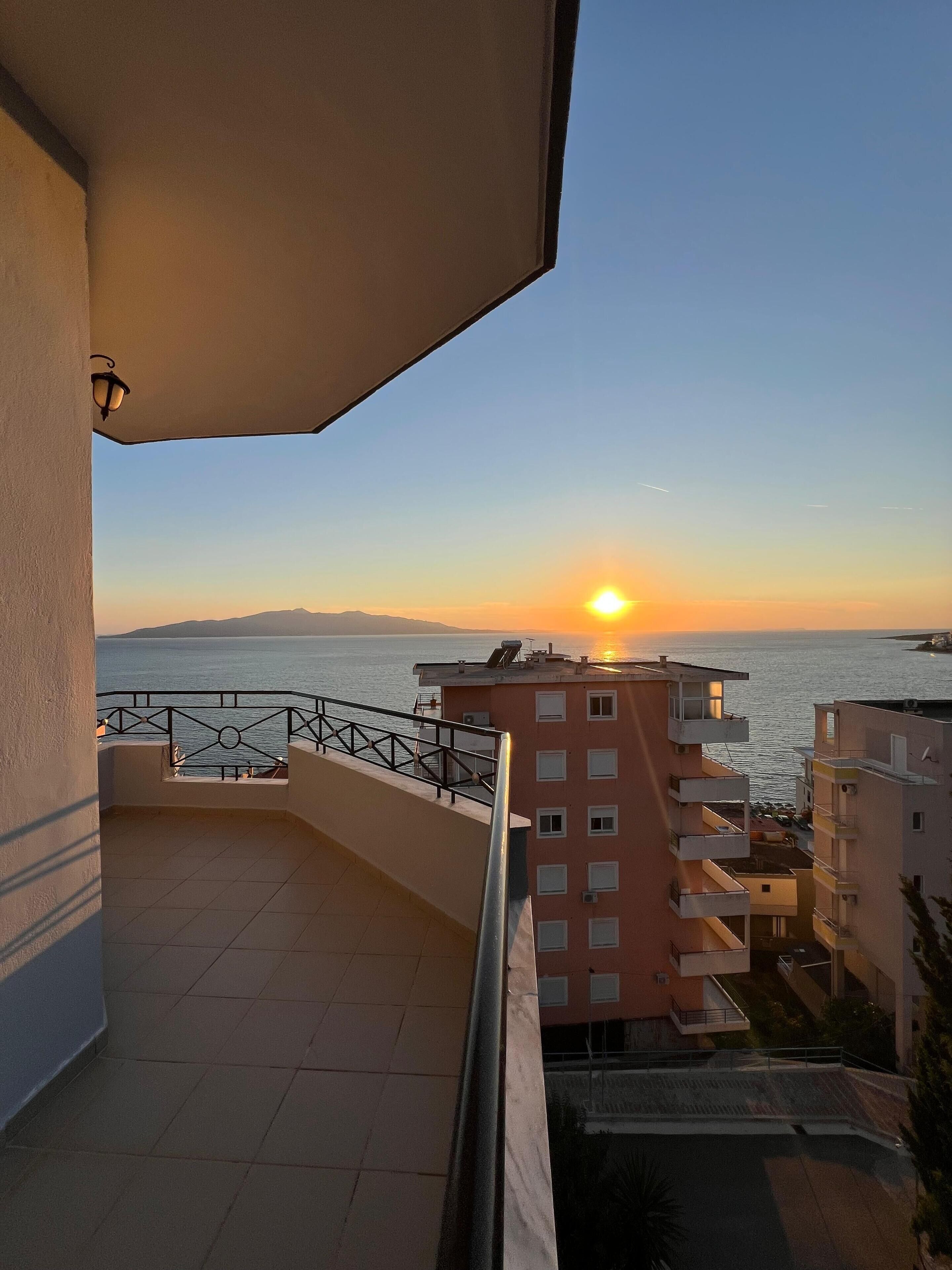 A balcony with a view of the sunset over the sea, adjacent buildings, and a tiled floor.