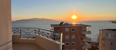 A balcony with a view of the sunset over the sea, adjacent buildings, and a tiled floor.