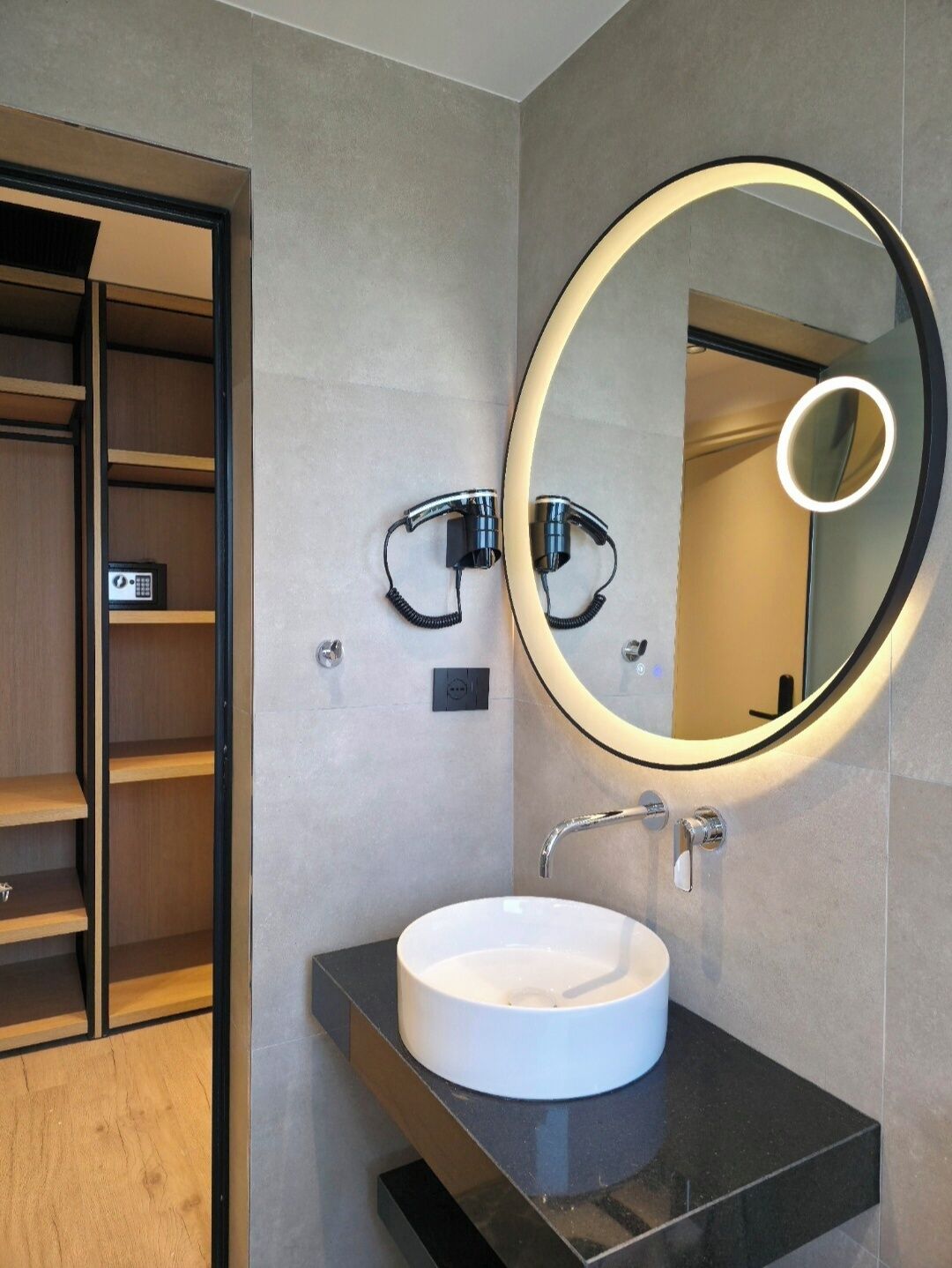 A modern bathroom with a round mirror, a white sink, and a wooden shelf.