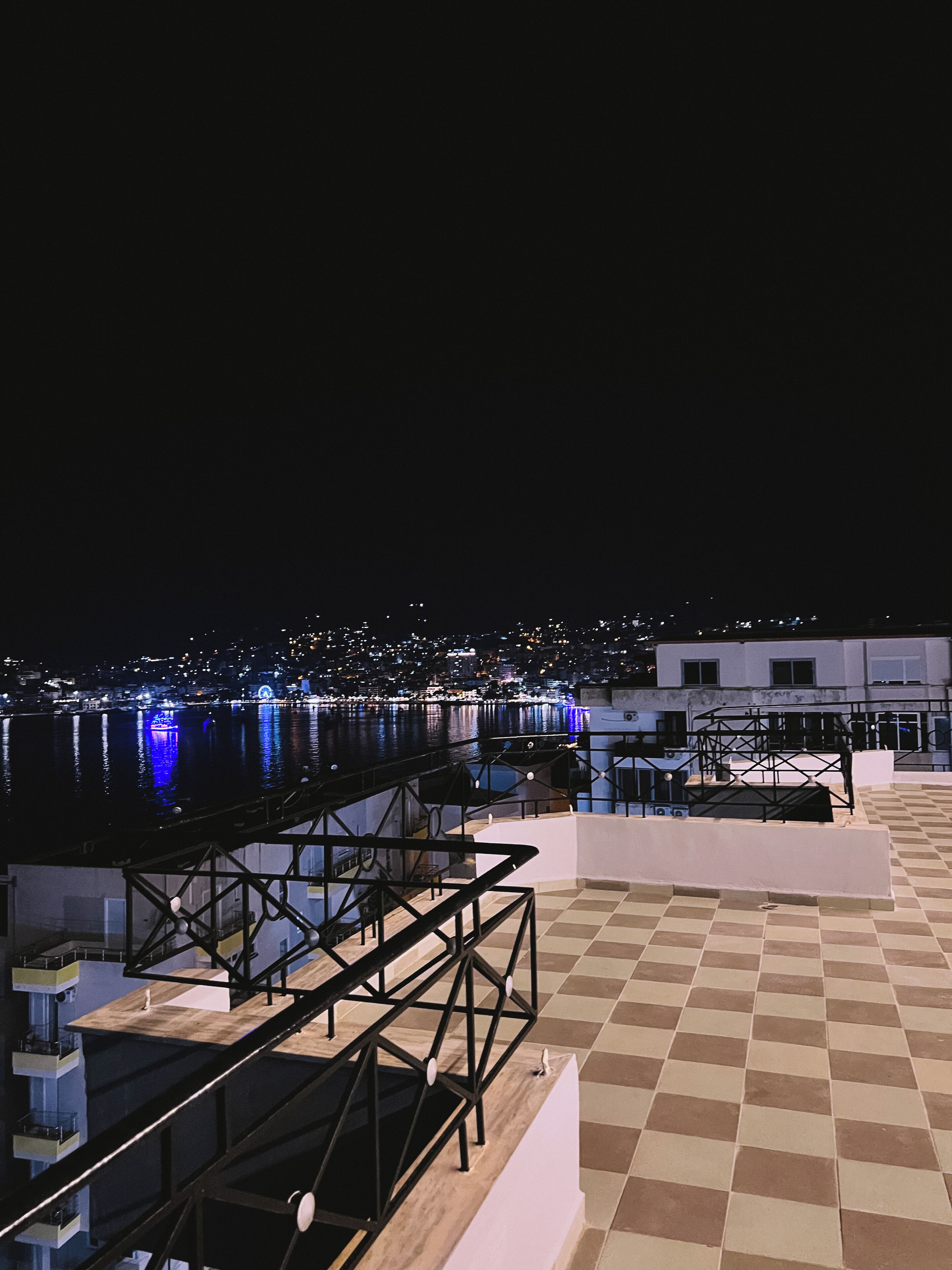 Una terraza en la azotea con suelo a cuadros, barandilla de metal y vista panorámica de la ciudad de noche.