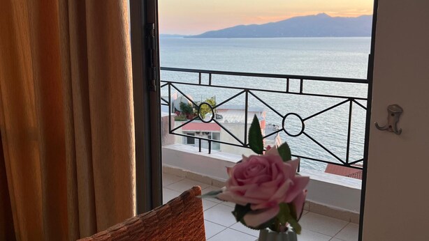 A room with a view of a body of water and mountains, a table with a vase of flowers, and a chair. - Blue Waves Hotel & Suites (Sarandë)