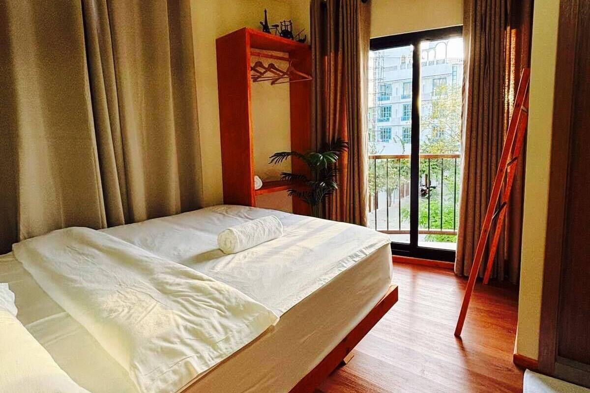 A hotel room with a bed, a wooden floor, a balcony with curtains, and a view of buildings outside.