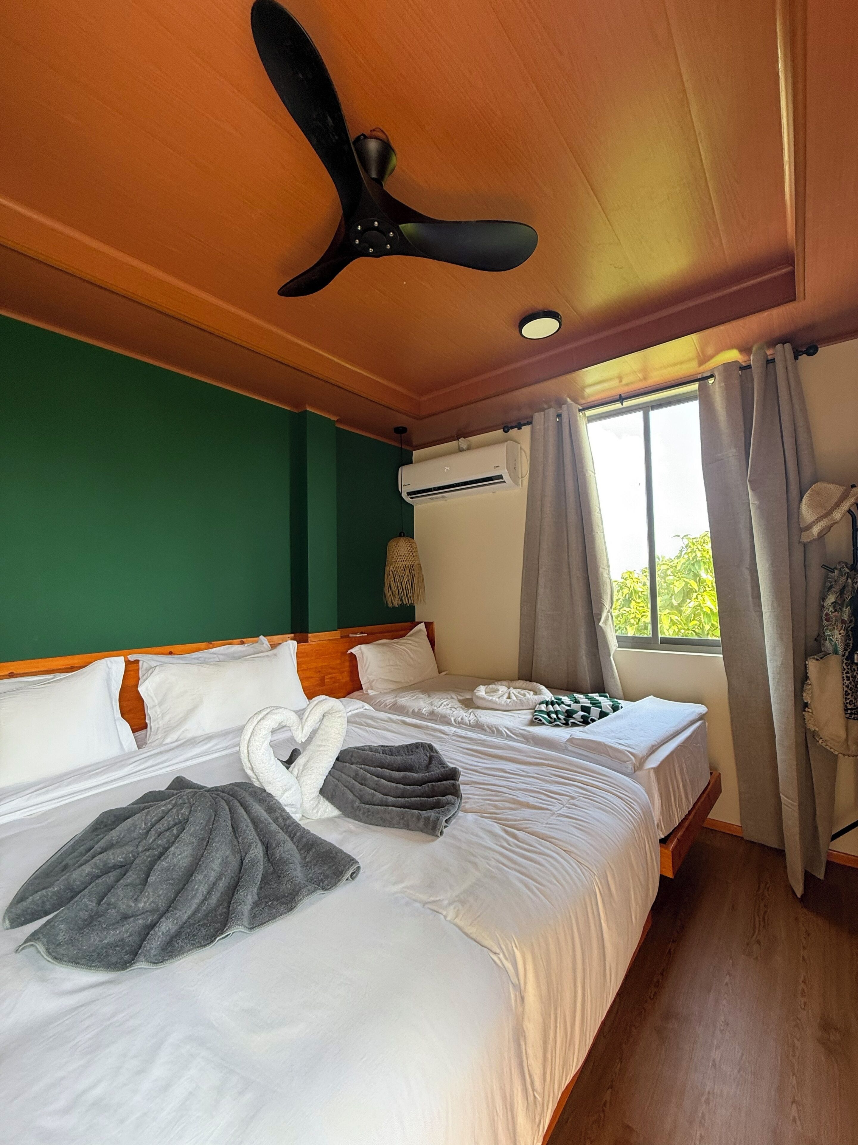 A hotel room with two beds, a ceiling fan, an air conditioning unit, and a view of greenery outside.