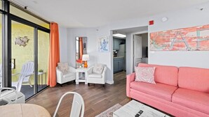 A living room with a pink sofa, white armchairs, a small table with a lamp, and a glass door leading to an outdoor area with a white chair. - Ocean View Corner King Suite Sleeps 4 (Myrtle Beach)