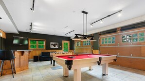 Een pooltafel met een rode viltbekleding, een tafelvoetbalspel, een bar met krukken, en een groot raam met uitzicht op groen.