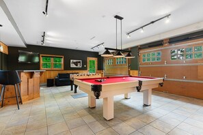 A pool table with a red felt surface, a foosball table, a bar area with stools, and a large window with a view of greenery.
