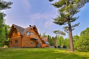 A log cabin with a balcony, a staircase, and a view of the surrounding forest.