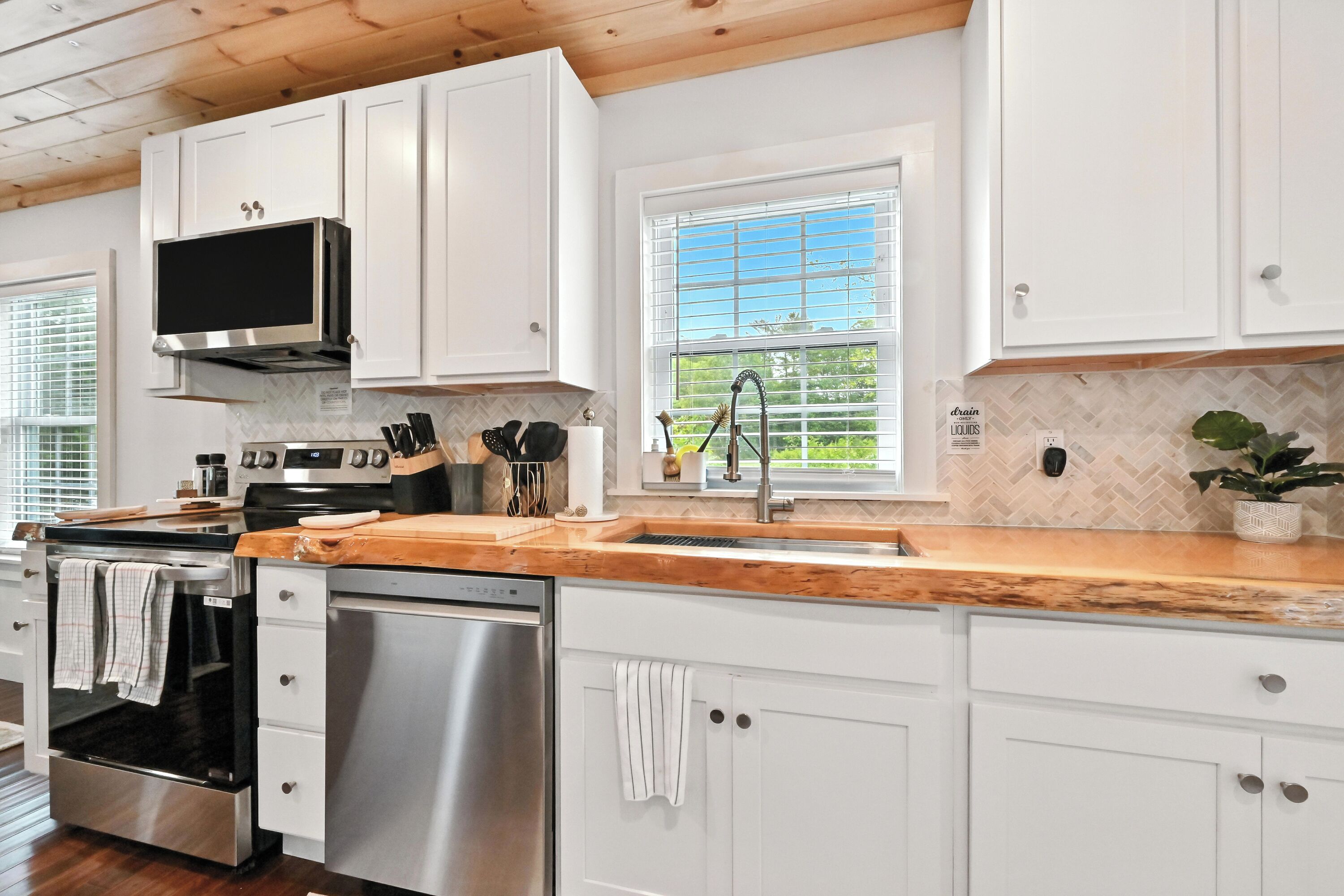 A modern kitchen with white cabinets, stainless steel appliances, a wooden countertop, and a window with blinds.