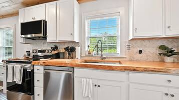 A modern kitchen with white cabinets, stainless steel appliances, a wooden countertop, and a window with blinds.