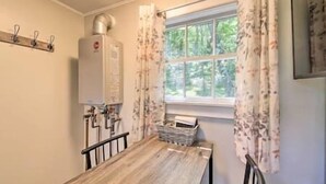 A dining area with a wooden table, chairs, a window with curtains, and a wall-mounted heater. - Happy Cottage Near Everything 11 (Edgecomb)