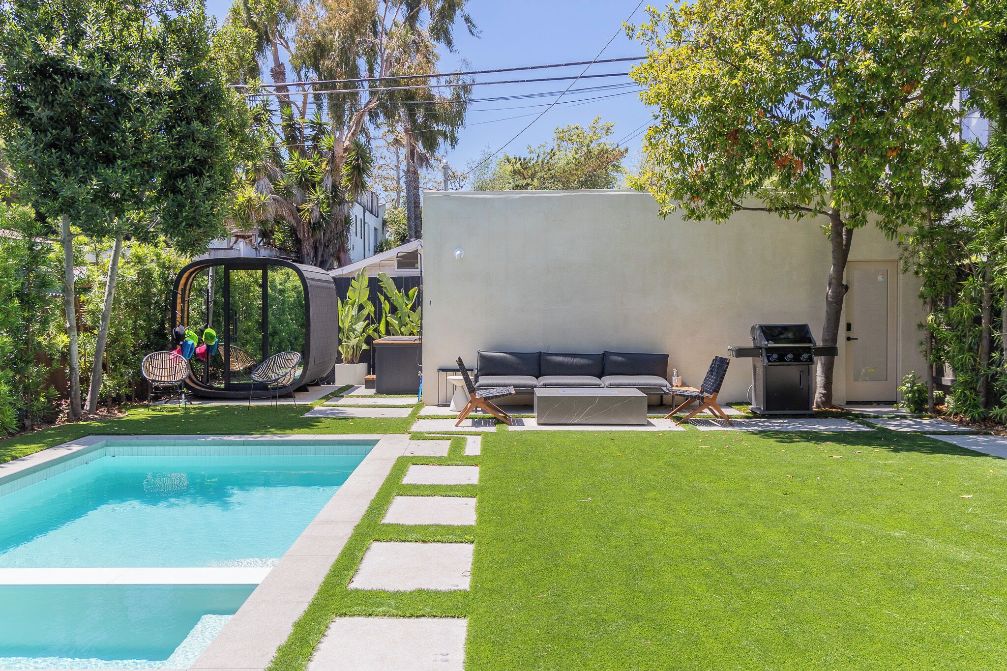 A backyard with a swimming pool, a covered seating area, a barbecue grill, and a lawn.
