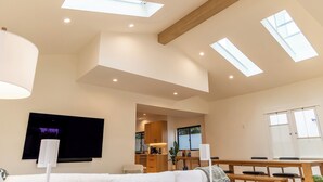 A modern living room with a flat-screen TV, white sofa, dining table, and skylights. - Private wellness home with pool, sauna, & cold plunge - in the center of Venice. (Los Angeles)