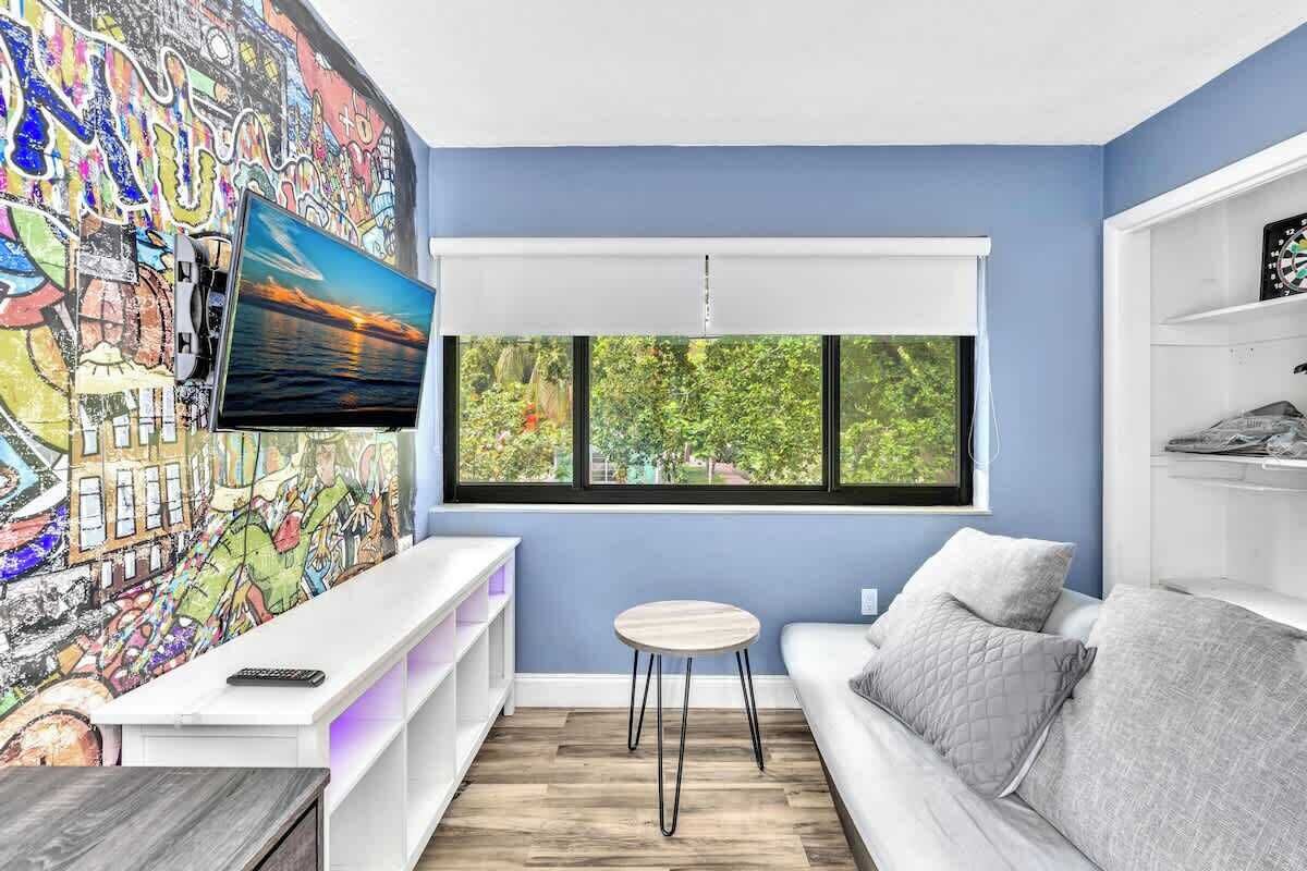 A modern living room with a large TV, a colorful mural, a window with a view of trees, and a white sofa.