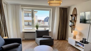 A modern living room with a grey sofa, a round coffee table, and a view of buildings through a window with curtains. - Seaside - Central 2-room app. for 2-4 pers. with balcony and WLAN (Sylt)