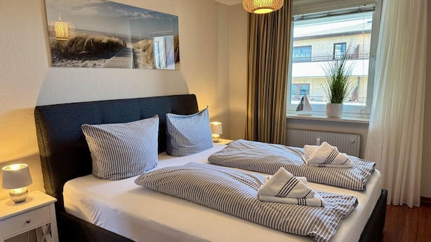A hotel room with a large bed, striped bedding, a nightstand with a lamp, a window with curtains, and a wall-mounted picture. - Seaside - Central 2-room app. for 2-4 pers. with balcony and WLAN (Sylt)