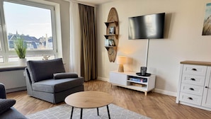 A modern living room with a grey sofa, a round coffee table, a TV on the wall, and a bookshelf. - Seaside - Central 2-room app. for 2-4 pers. with balcony and WLAN (Sylt)