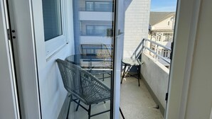 A balcony with a glass door, a round table, two wicker chairs, and a view of a building with multiple windows. - Seaside - Central 2-room app. for 2-4 pers. with balcony and WLAN (Sylt)