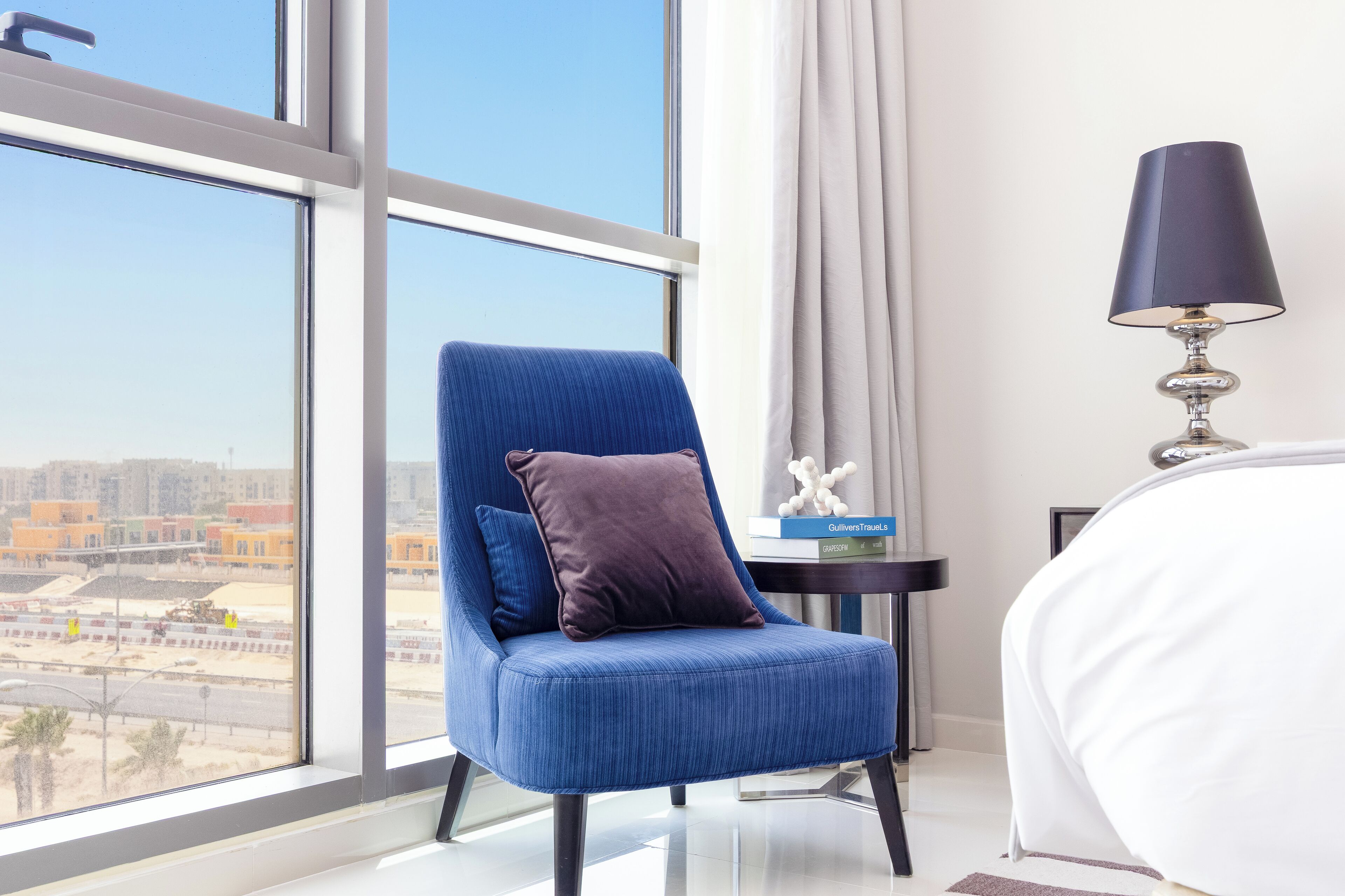 A hotel room with a large window, a blue upholstered chair with a dark cushion, a bedside table with a lamp, and a bed with white bedding.