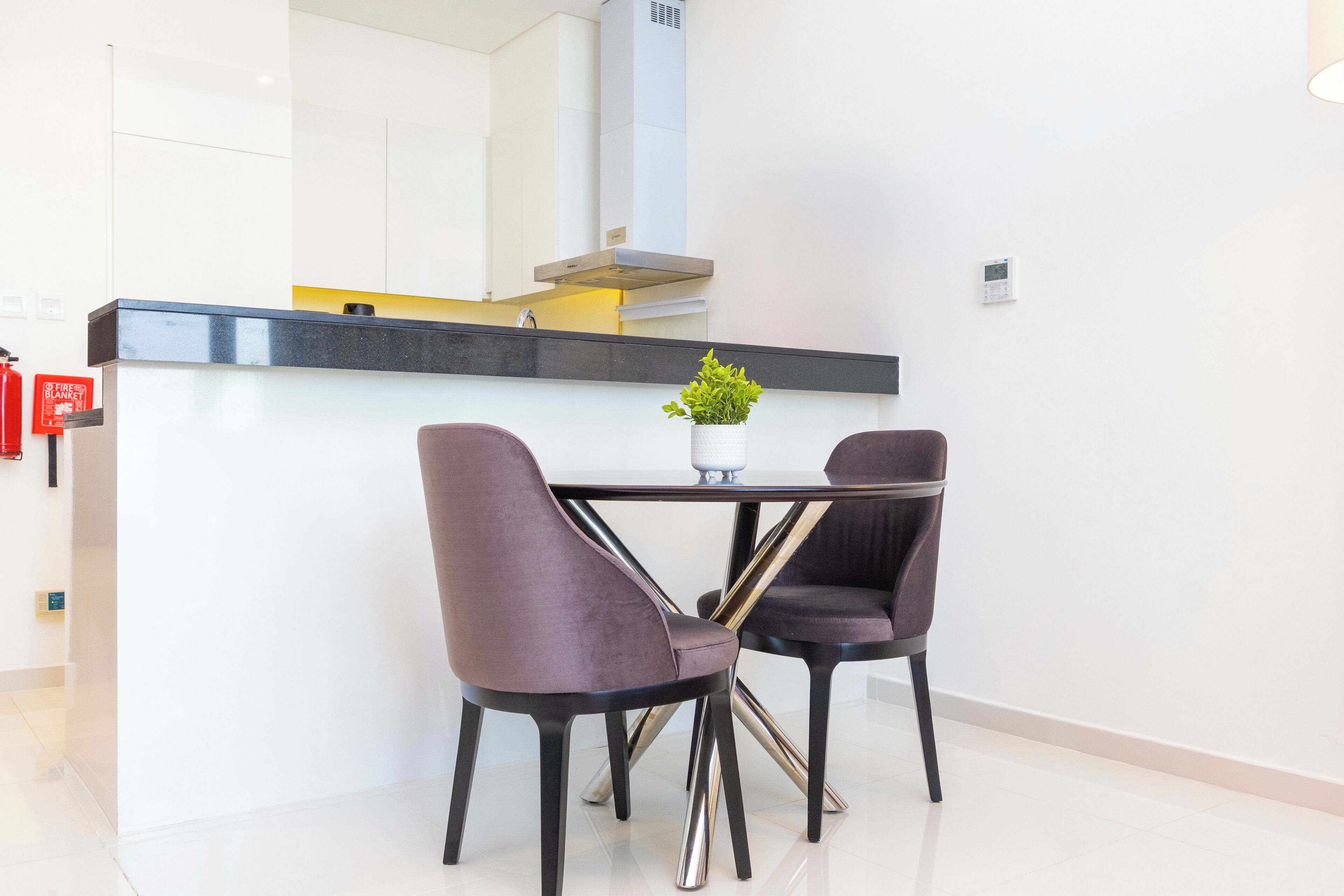 Een moderne keuken met een zwart aanrecht, roestvrijstalen apparatuur en twee paarse stoelen rondom een kleine tafel met een plant in pot.
