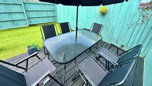 A deck with a glass-top table and four chairs, surrounded by a green fence and a house in the background.