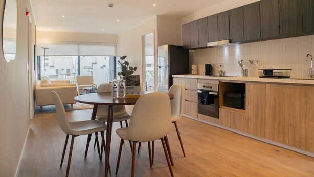 Una cocina moderna con armarios de madera, un comedor con mesa y sillas redondas, y vistas de la ciudad a través de grandes ventanales.