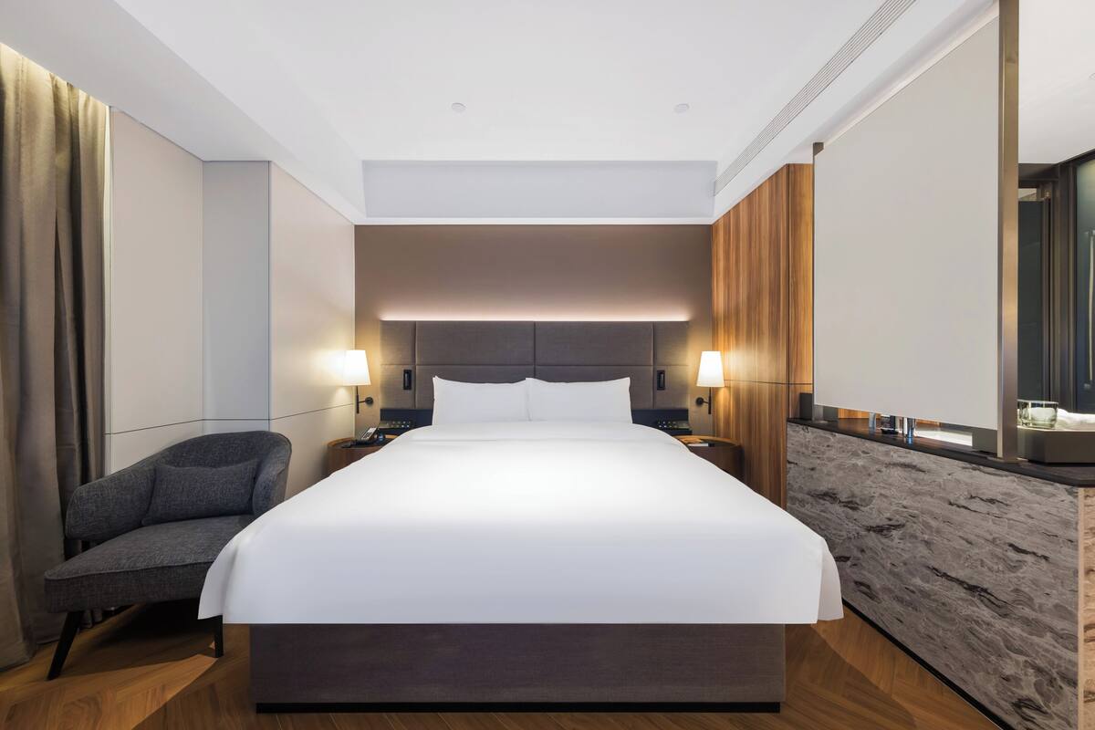 Une chambre d’hôtel moderne avec un grand lit, deux tables de chevet avec des lampes, un fauteuil gris et un mur décoratif en bois.