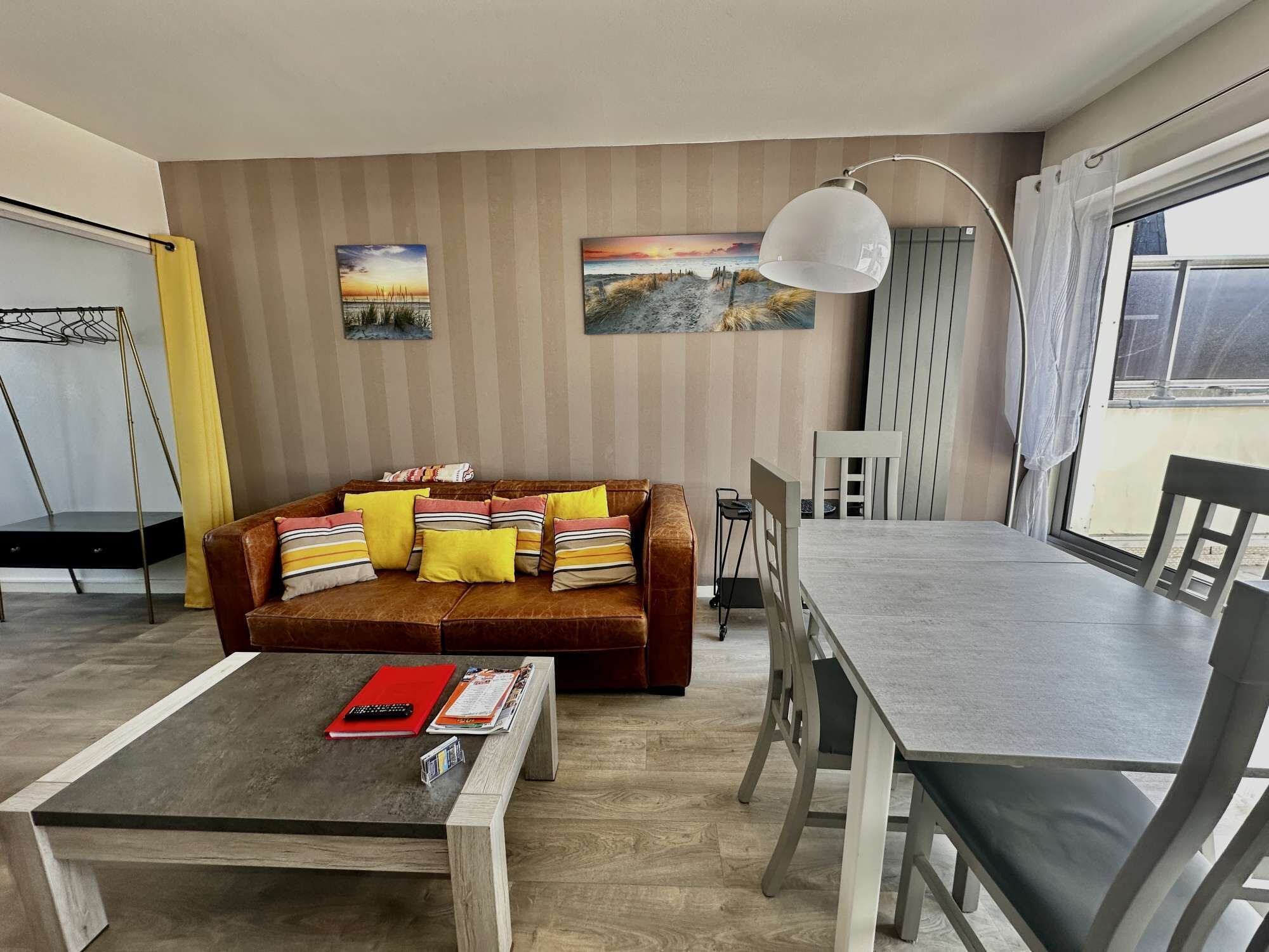 Ein modernes Wohnzimmer mit einer Couch, einem Couchtisch, einem Esstisch und Stühlen. An den Wänden hängen zwei Gemälde, eine Pendelleuchte und ein Fenster mit Vorhängen.