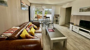 A modern living room with a brown leather sofa, dining table, and chairs, a TV on a wooden cabinet, and a view of the outdoors through large windows. - La mouette rieuse, terrace, sea view, beach access (Bernières-sur-Mer)