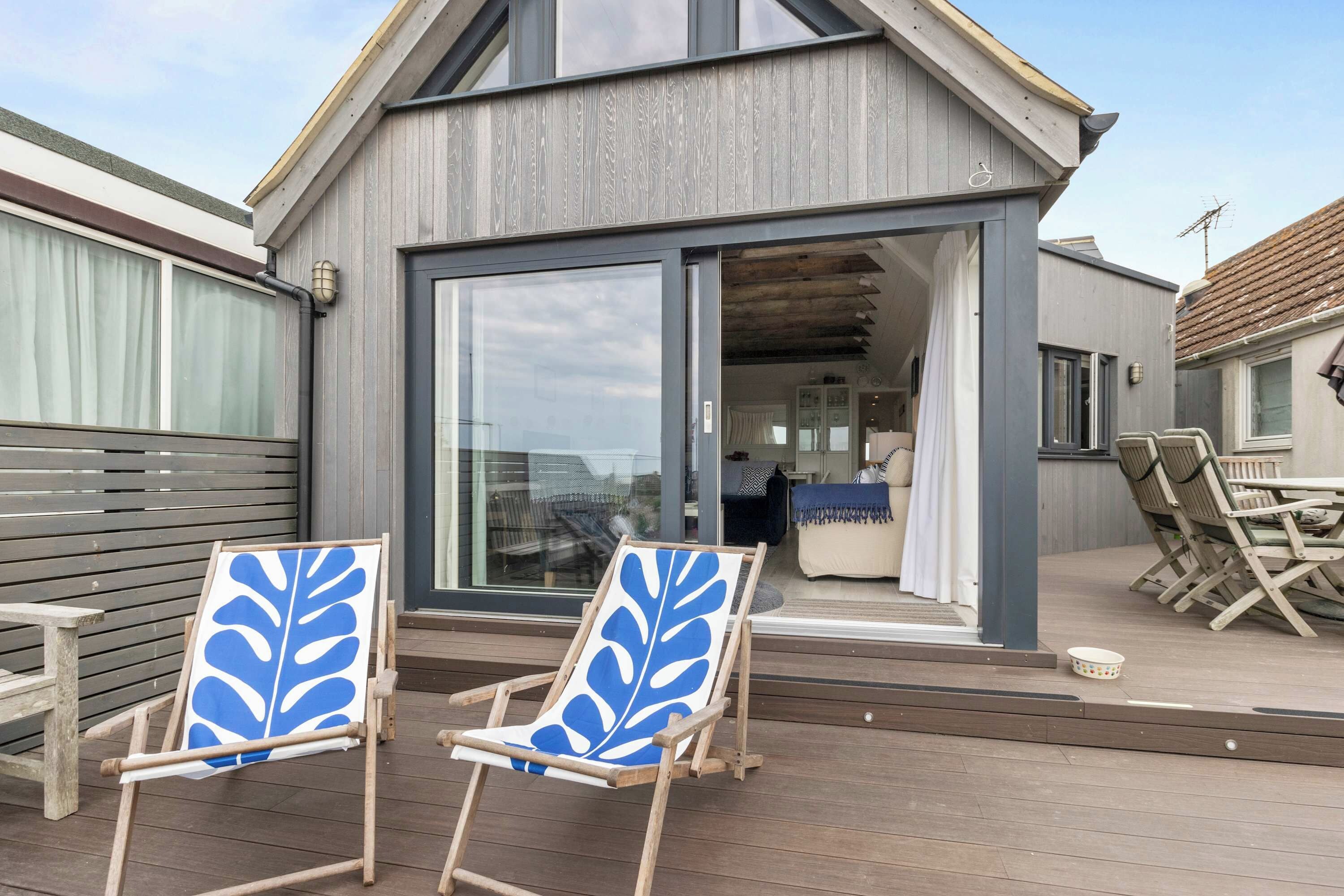 Una casa moderna con un amplio ventanal que da a una sala de estar, dos sillas de madera con motivos de hojas azules y una terraza de madera.