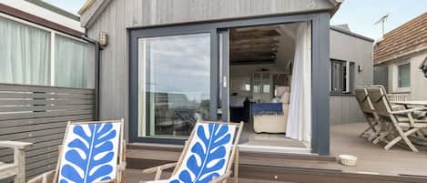 Una casa moderna con un amplio ventanal que da a una sala de estar, dos sillas de madera con motivos de hojas azules y una terraza de madera.