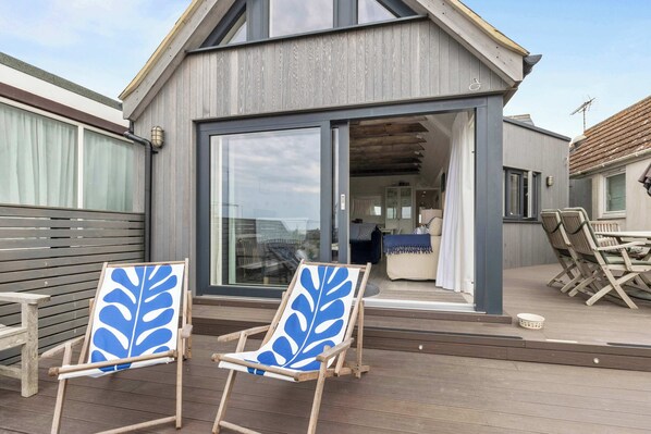 Ein modernes Haus mit einer großen Glasdoor, die zu einem Wohnbereich führt, zwei Holzliegen mit blattförmigen blauen Mustern und eine Holzterrasse.