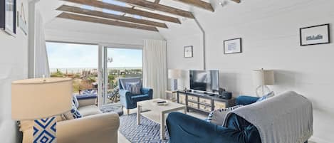 Amplia sala de estar con un televisor grande, muebles azules y vista a la playa a través de puertas correderas de vidrio.