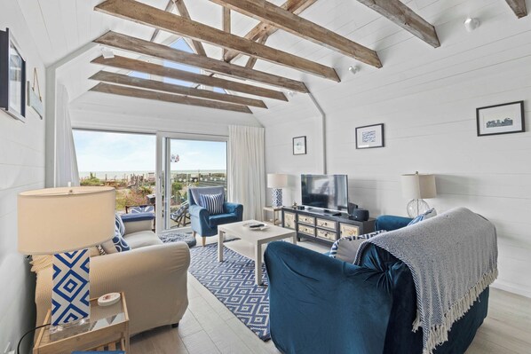A spacious living room with a large TV, blue furniture, and a view of the beach through the sliding glass doors.