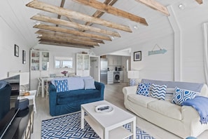 A spacious living area with a high ceiling and exposed beams, a kitchen in the background, and a sign indicating a beach location.